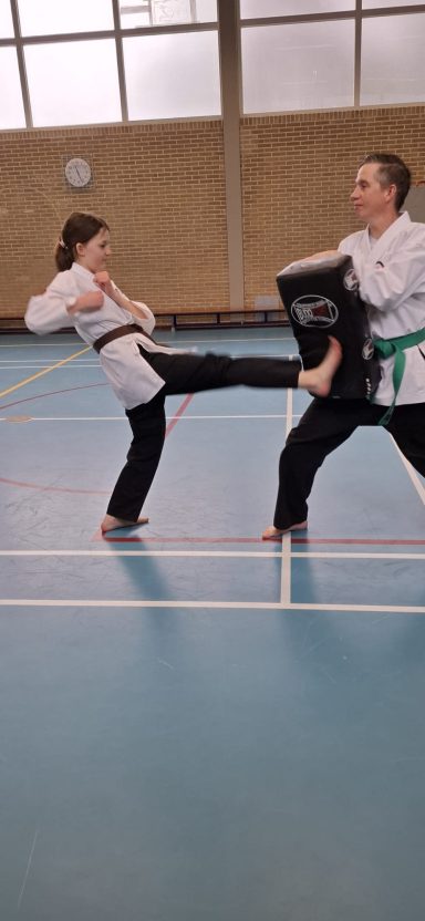 Twee martial arts beoefenaars in een trainingsruimte, één trapt tegen een schild.